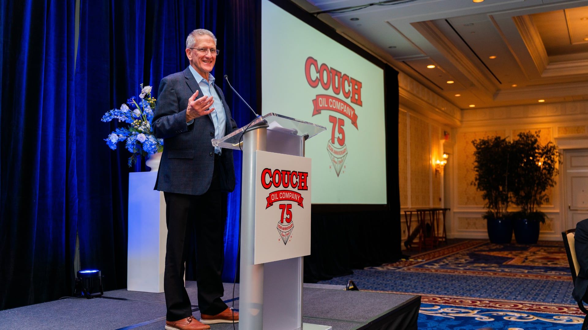 Speaker at a podium during a Couch Oil Company event, with the company’s 75th-anniversary logo on screen